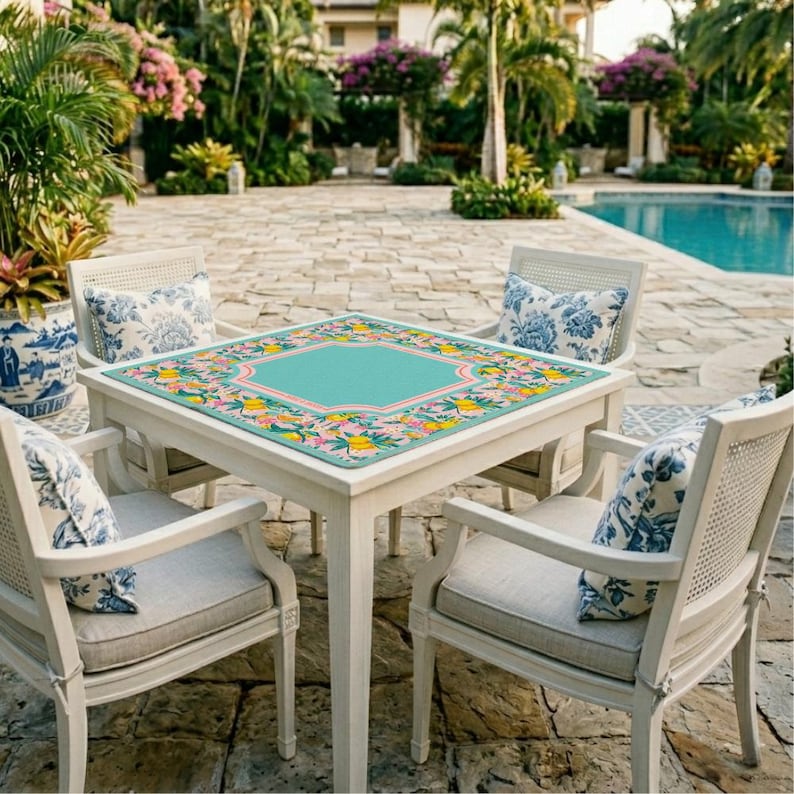 May include: A square table with a light-colored frame and a decorative mat featuring a teal center, a pink border, and a colorful pattern of yellow lemons and green leaves. Four chairs with light-colored frames and blue and white floral pillows.