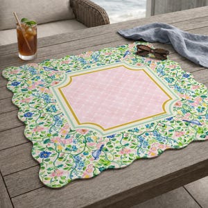 May include: A square placemat with scalloped edges featuring a central pink diamond-patterned panel framed by gold and light blue borders. The surrounding area displays a botanical print of blue birds, pink flowers, and green vines on a white background.
