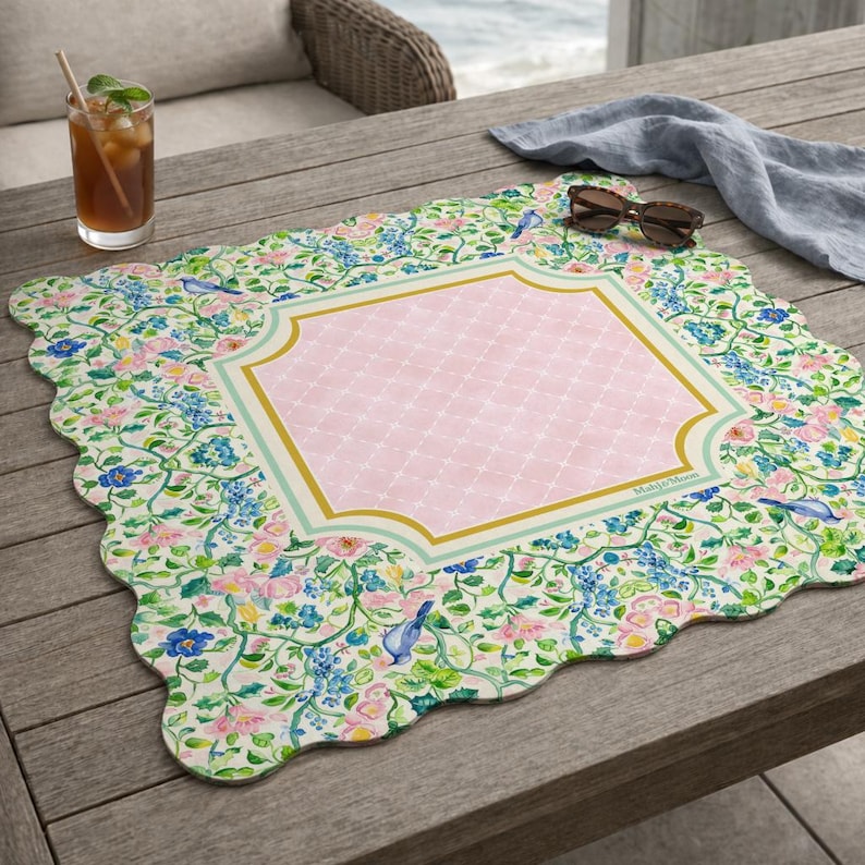 May include: A square placemat with scalloped edges featuring a central pink diamond-patterned panel framed by gold and light blue borders. The surrounding area displays a botanical print of blue birds, pink flowers, and green vines on a white background.