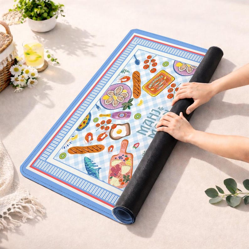 May include: A rectangular mat with a light blue and white gingham pattern, featuring illustrations of food items like oysters, bread, and a tin labeled ANCHOVIES. The mat has a blue, white, and red striped border and is partially rolled up.