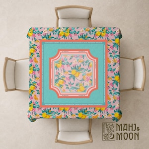 May include: A square tablecloth with a pink background and a pattern of yellow lemons, orange slices, and white flowers. The center features a turquoise frame with a floral design. The tablecloth is set on a square table with four chairs.