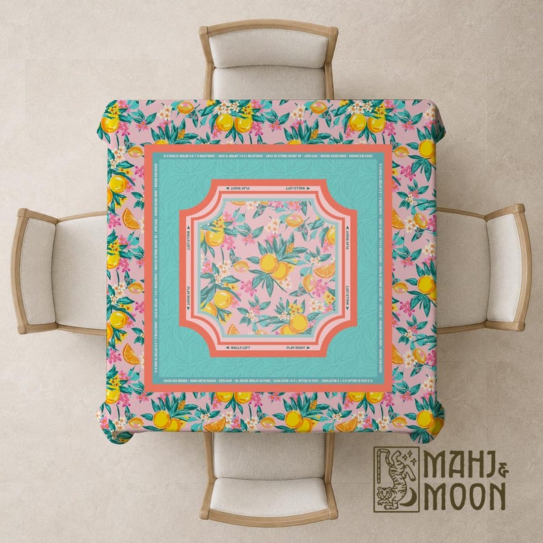 May include: A square tablecloth with a pink background and a pattern of yellow lemons, orange slices, and white flowers. The center features a turquoise frame with a floral design. The tablecloth is set on a square table with four chairs.