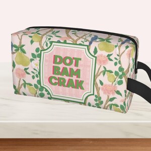 May include: A rectangular wash bag with a floral design of pears, flowers, and birds. It has a black zip and handle. The centre of the bag features the words "DOT BAM CRAK" in green and pink.