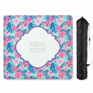 May include: A rectangular beach blanket with a vibrant floral pattern in pink, blue, and green. The centre features a white quatrefoil shape with the text "MAHJ & MOON". A black carrying bag is included.