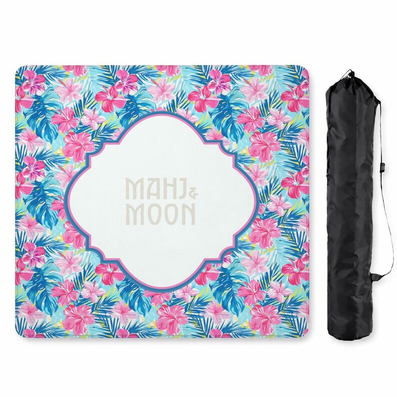May include: A rectangular beach blanket with a vibrant floral pattern in pink, blue, and green. The centre features a white quatrefoil shape with the text "MAHJ & MOON". A black carrying bag is included.
