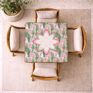 Op de afbeelding: Een vierkante tafel met een feestelijk patroon van roze huizen, groene dennenbomen en sneeuwpoppen, in het midden voorzien van een groot, gelaagd bloemmotief in tinten roze, wit en saliegroen, omringd door vier stoelen met lichtbeige kussens.