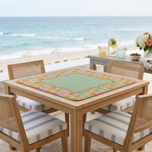May include: A square table mat with a light green center and a border featuring a colorful pattern of small orange, yellow, and blue sea turtles. The mat is placed on a wooden table surrounded by four chairs with white and blue striped cushions.
