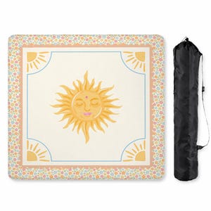 Puede incluir: Una manta de picnic crema con una gran ilustraci&oacute;n de un sol sonriente en el centro. El sol tiene los ojos cerrados y una boca rosa. La manta tiene un borde floral y ribete azul. Una bolsa de transporte negra est&aacute; a la derecha. Aproximadamente 150 cm x 150 cm.