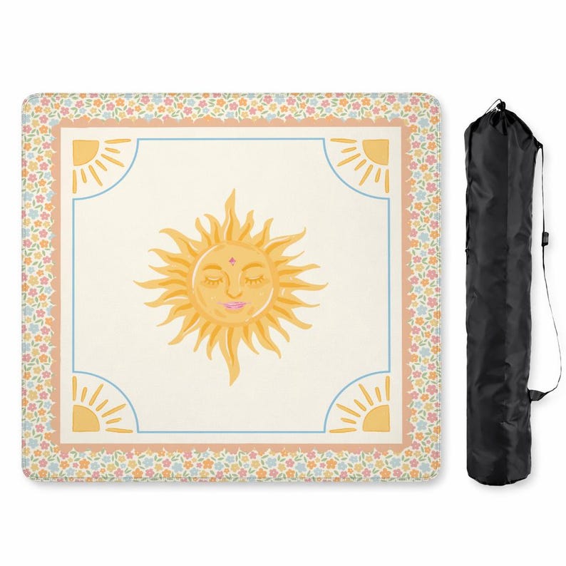Puede incluir: Una manta de picnic crema con una gran ilustraci&oacute;n de un sol sonriente en el centro. El sol tiene los ojos cerrados y una boca rosa. La manta tiene un borde floral y ribete azul. Una bolsa de transporte negra est&aacute; a la derecha. Aproximadamente 150 cm x 150 cm.