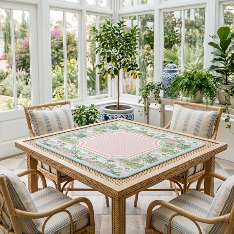 Op de afbeelding: Een vierkante rotan tafel met vier bijpassende stoelen, elk met gestreepte kussens. De tafel is bedekt met een roze en groen patroon. De setting is in een serre met grote ramen en potplanten.