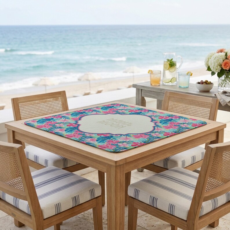 May include: A wooden table with a floral tablecloth, set for outdoor dining. The tablecloth features pink and blue flowers with green leaves. The table is surrounded by four chairs with striped cushions. The sea is visible in the background.