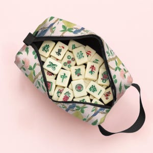 May include: A floral print zipped pouch filled with ivory Mahjong tiles. The pouch has a black handle and a pattern of pink flowers, green leaves, and blue birds. The tiles have red, green, and black symbols.