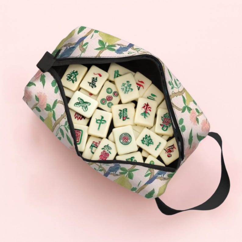 May include: A floral print zipped pouch filled with ivory Mahjong tiles. The pouch has a black handle and a pattern of pink flowers, green leaves, and blue birds. The tiles have red, green, and black symbols.