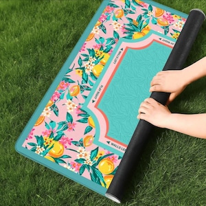 May include: A rectangular playmat featuring a pink background with a pattern of yellow lemons, green leaves, and white flowers. The center has a teal section with text reading WALLS LEFT, WALLS RIGHT, PLAY LEFT, and PLAY RIGHT in black.