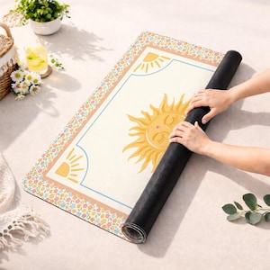 Puede incluir: Una esterilla de yoga enrollada con una superficie crema, con un gran sol amarillo con los ojos cerrados. La esterilla tiene un borde floral y un marco azul. La esterilla se est&aacute; enrollando. Las dimensiones de la esterilla son en cent&iacute;metros.
