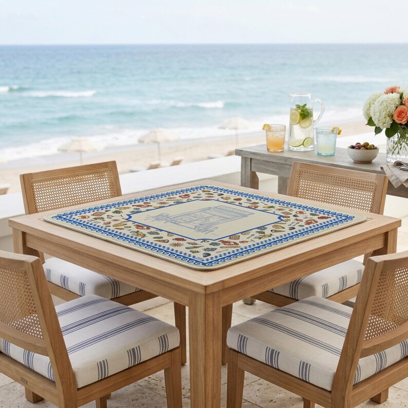 Puede incluir: Un mantel individual cuadrado con una ilustraci&oacute;n central del Arco del Triunfo rodeada por un borde de iconos coloridos de comida y bebida mediterr&aacute;nea, enmarcado por un patr&oacute;n geom&eacute;trico azul y blanco sobre un fondo beige claro.
