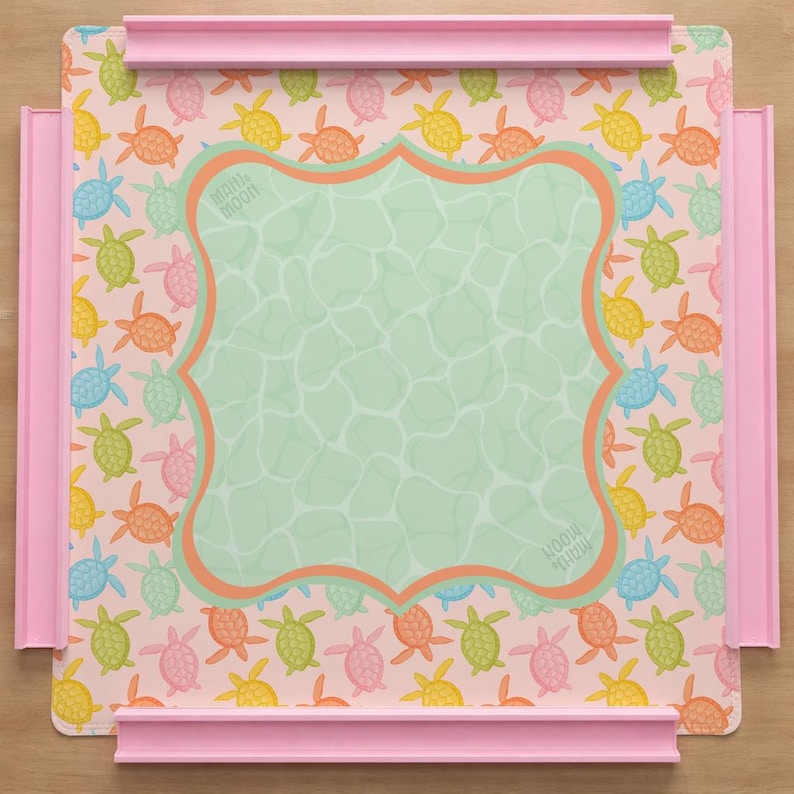 May include: A square puzzle board with a light pink background featuring a repeating pattern of small colorful turtles in yellow, orange, green, and blue. A central light green scalloped frame has a water-like texture and the text Mahi Moon.