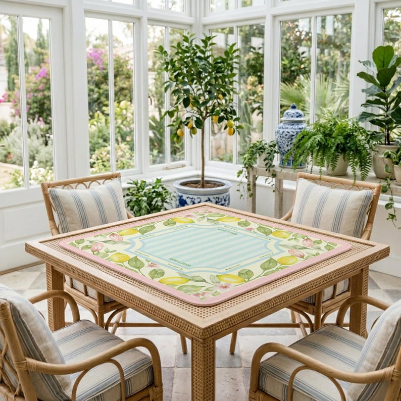 K&ouml;nnte beinhalten: Ein quadratischer Rattantisch mit vier passenden St&uuml;hlen, jeweils mit gestreiften Kissen. Der Tisch hat eine dekorative Matte mit hellblauem Mittelteil, rosa Rand und Zitronen- und Blumendesign. Die Szene ist in einem Wintergarten mit einem Zitronenbaum.