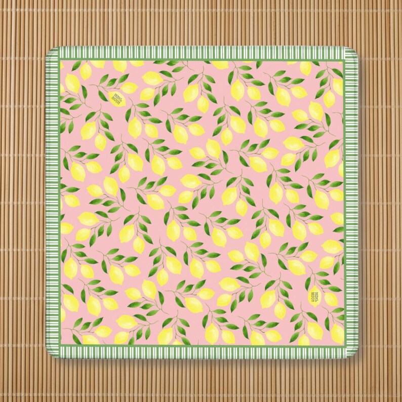 May include: A square fabric napkin featuring a repeating pattern of yellow lemons and green leaves on a light pink background, framed by a green and white striped border. The fabric has a small rectangular tag with text on the surface.