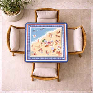 May include: Overhead view of a square table with a beach scene illustration, surrounded by four wicker chairs with cream-colored cushions. The table has a blue border and a detailed beach illustration with people, umbrellas, and the ocean. A potted plant sits in the upper left corner.