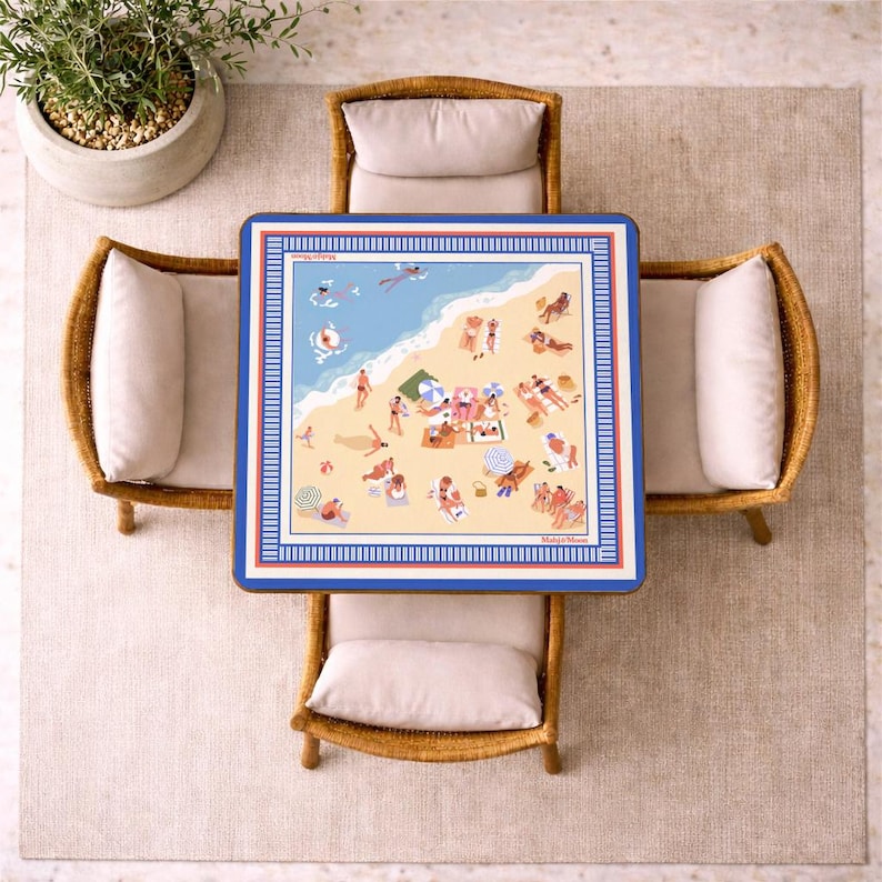 May include: Overhead view of a square table with a beach scene illustration, surrounded by four wicker chairs with cream-colored cushions. The table has a blue border and a detailed beach illustration with people, umbrellas, and the ocean. A potted plant sits in the upper left corner.