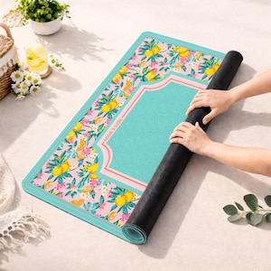 May include: A rectangular yoga mat featuring a pink background with a repeating pattern of yellow lemons, green leaves, and small white flowers. The mat has a solid teal central panel with a thin pink border and the text "MINI & MOON" printed on it.