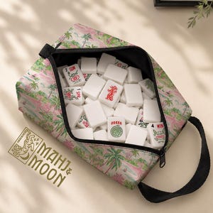 Op de afbeelding: Een tas met ritssluiting gevuld met witte Mahjong-tegels. De tas heeft een tropische print met palmbomen en roze accenten. De tas heeft een zwarte handgreep en de tekst "MAHJ & MOON" is zichtbaar.