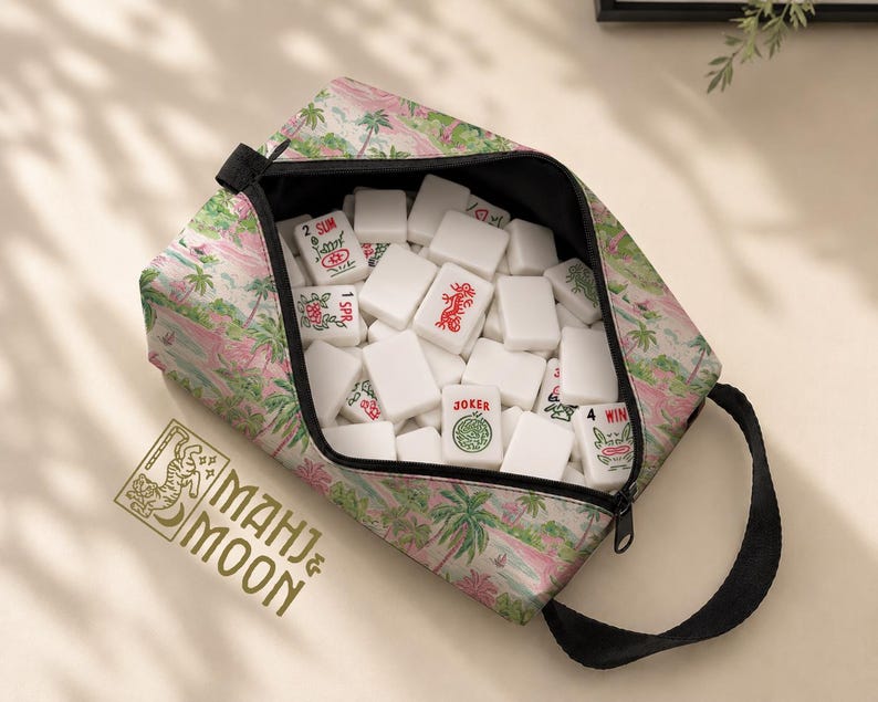 Op de afbeelding: Een tas met ritssluiting gevuld met witte Mahjong-tegels. De tas heeft een tropische print met palmbomen en roze accenten. De tas heeft een zwarte handgreep en de tekst "MAHJ & MOON" is zichtbaar.