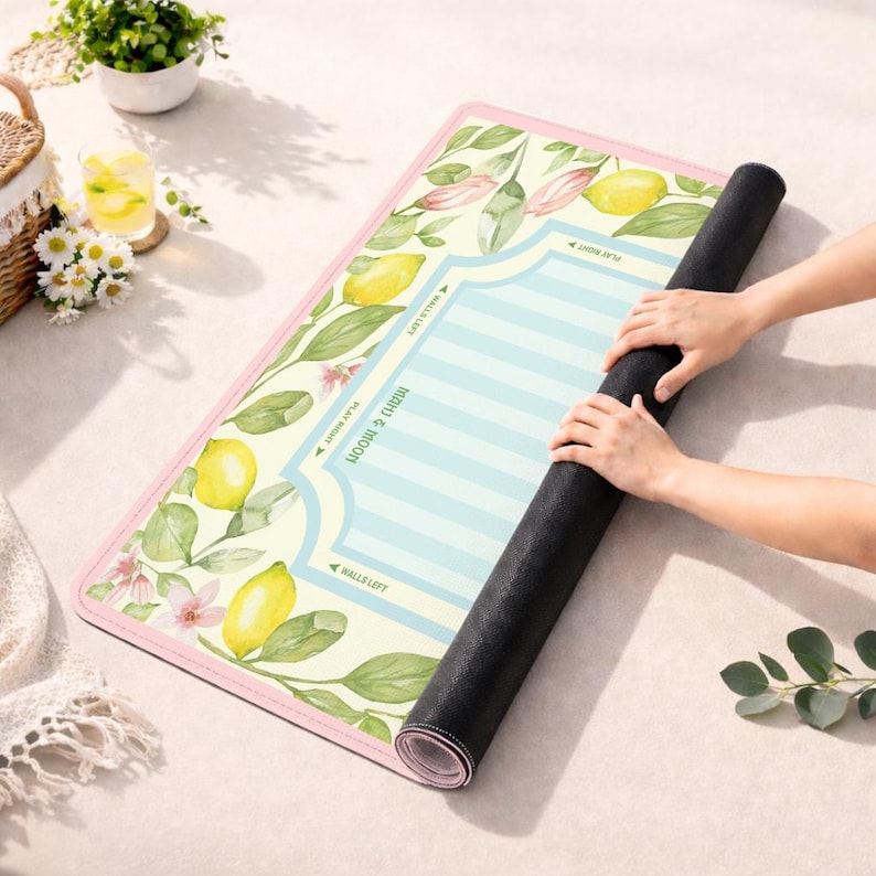 K&ouml;nnte beinhalten: Eine zusammengerollte Yogamatte mit einem Zitronen- und Blumenmuster. Die Matte hat einen hellrosa Rand und einen Mittelteil mit blauen Streifen und Text. Das Design umfasst gelbe Zitronen, gr&uuml;ne Bl&auml;tter und rosa Bl&uuml;ten.