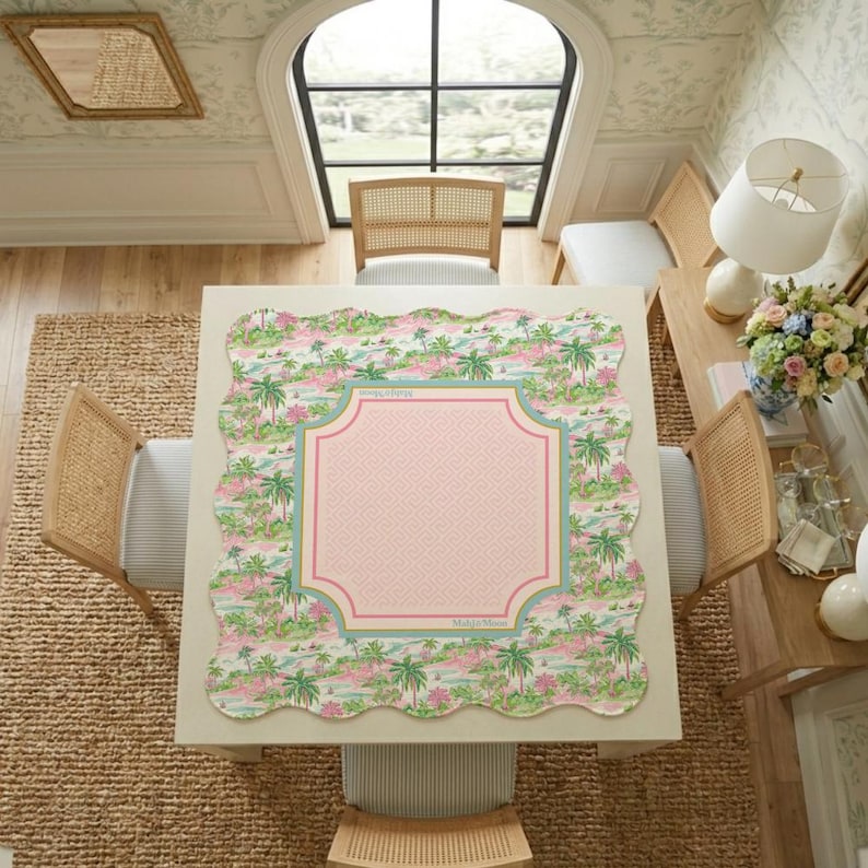 May include: A square tablecloth with scalloped edges featuring a tropical palm tree and landscape print in shades of pink and green. The centre has a solid light pink panel with a geometric border in pink and teal, labelled with the text Mally's Mom.