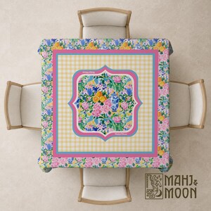 May include: A square tablecloth featuring a central panel with a floral and citrus pattern, surrounded by a yellow gingham border and a pink frame. The outer edge displays a repeating print of blue and white vases, oranges, and pink flowers.