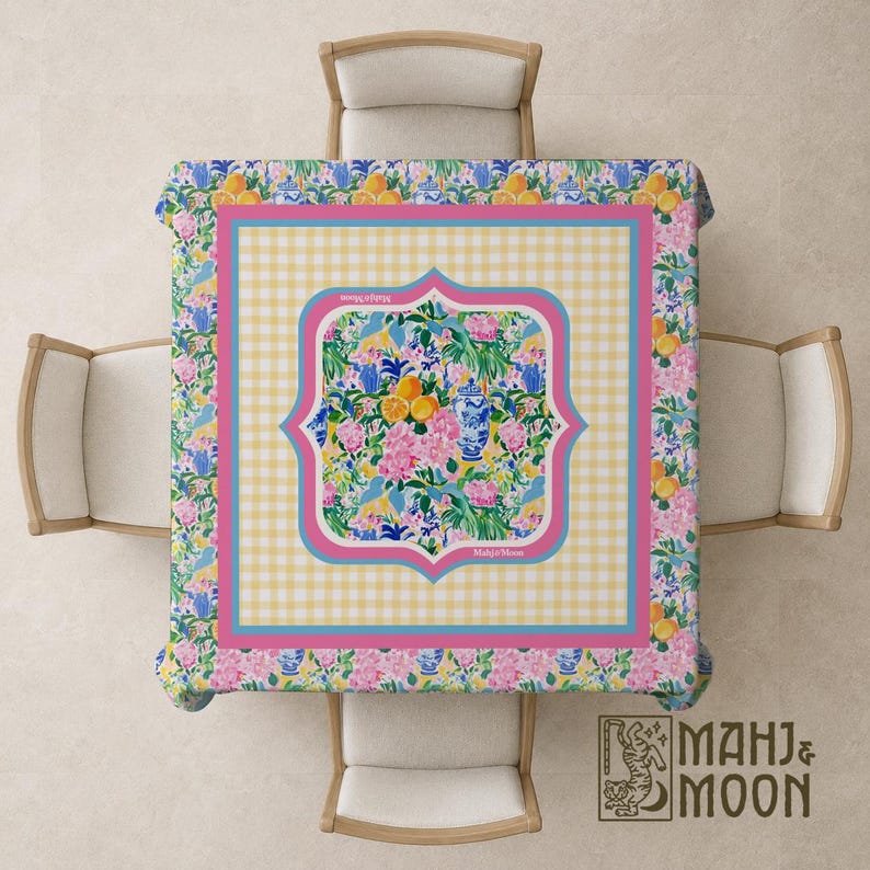 May include: A square tablecloth featuring a central panel with a floral and citrus pattern, surrounded by a yellow gingham border and a pink frame. The outer edge displays a repeating print of blue and white vases, oranges, and pink flowers.