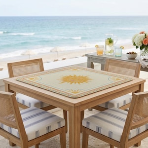 Puede incluir: Una mesa cuadrada de madera con cuatro sillas, situada en un patio con vistas al mar. La mesa tiene un mantel individual crema con un dise&ntilde;o de sol amarillo. Las sillas tienen respaldos tejidos y cojines a rayas. Hay bebidas y flores en una mesa auxiliar.