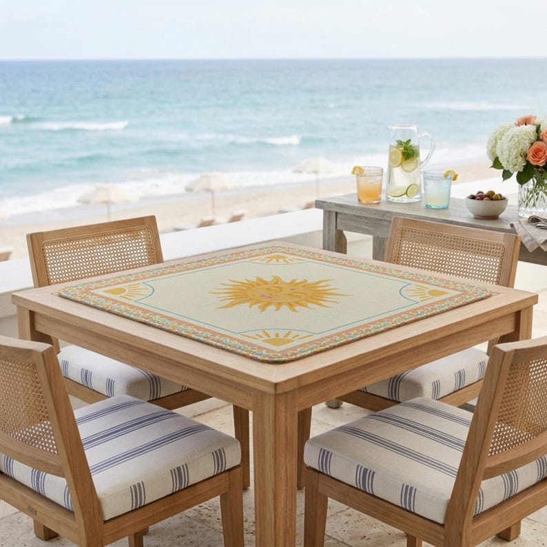 Puede incluir: Una mesa cuadrada de madera con cuatro sillas, situada en un patio con vistas al mar. La mesa tiene un mantel individual crema con un dise&ntilde;o de sol amarillo. Las sillas tienen respaldos tejidos y cojines a rayas. Hay bebidas y flores en una mesa auxiliar.