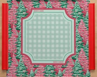 May include: A square game mat featuring a red background with a repeating green and white pine tree pattern. The center has a light green and white checkered square with a dark pink border and the text MAHJ & MOON. Four red racks frame the mat.