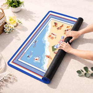 May include: A rectangular beach-themed mat with a blue border and a detailed illustration of a beach scene. The mat features people sunbathing, swimming, and relaxing on the sand. The mat is being rolled up, revealing its black underside.