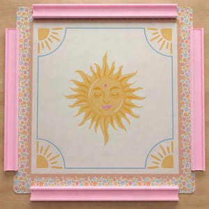 Puede incluir: Un tablero de Scrabble decorativo con una ilustraci&oacute;n central de un sol con los ojos cerrados y labios rosas. El tablero tiene un borde floral y un marco rosa, con dise&ntilde;os de sol en las esquinas. El tablero est&aacute; dise&ntilde;ado para jugar al juego de palabras.
