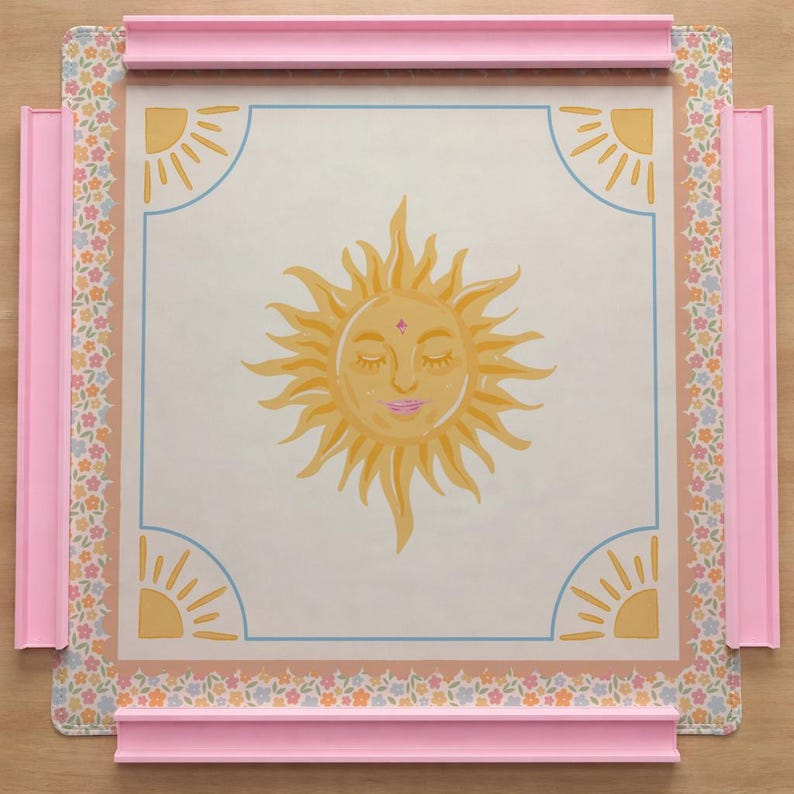 Puede incluir: Un tablero de Scrabble decorativo con una ilustraci&oacute;n central de un sol con los ojos cerrados y labios rosas. El tablero tiene un borde floral y un marco rosa, con dise&ntilde;os de sol en las esquinas. El tablero est&aacute; dise&ntilde;ado para jugar al juego de palabras.