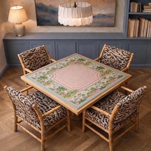 Op de afbeelding: Een vierkante eettafel met een roze en groen bloemenontwerp, omringd door vier stoelen met bamboe frames en luipaardprint bekleding. De tafel staat in een kamer met een blauwe muur en een decoratieve lamp.