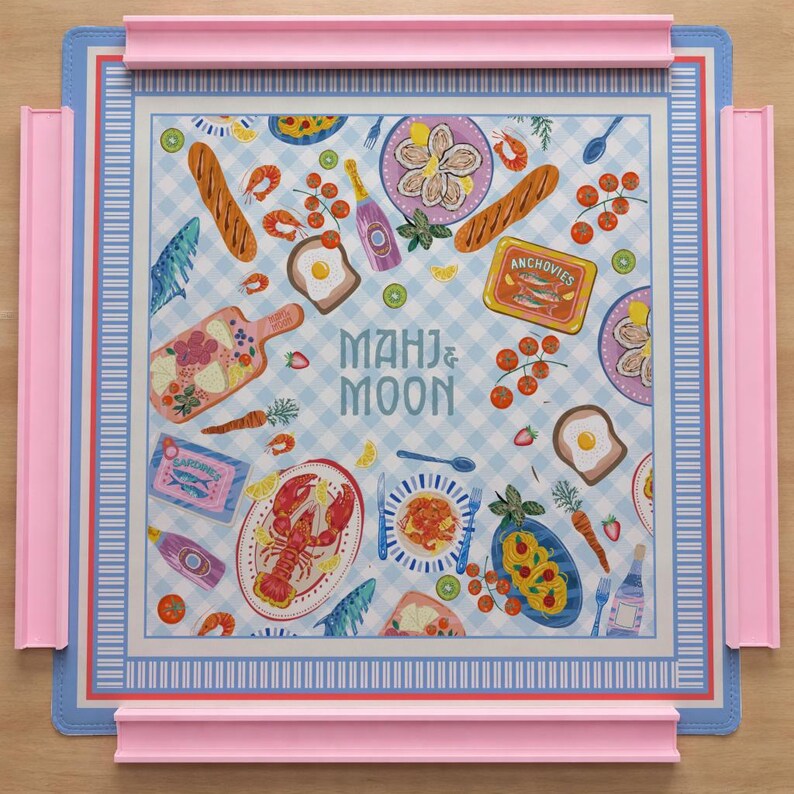 May include: A square Mahj & Moon game mat featuring a light blue and white checkered pattern decorated with illustrations of food items like lobster, bread, anchovies, and sardines, framed by a blue border with four attached pink tile racks.
