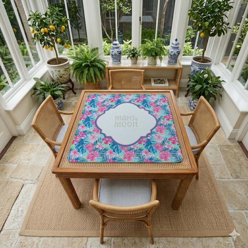 May include: A square wooden table with a floral mat featuring pink and blue flowers. Four wicker chairs surround the table. The mat has the text "MAHI & MOON" in the centre. The room has large windows and potted plants, creating a bright space.