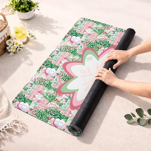 Op de afbeelding: Een rechthoekige yogamat met een herhalend patroon van roze huizen, groene dennenbomen en sneeuwpoppen. De mat heeft een grote, centrale, meerlaagse bloemachtige afbeelding in roze, wit en groen met de tekst Mahi Moon erop gedrukt.