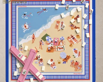 May include: A square Mahj & Moon game mat featuring a beach scene with people swimming and sunbathing. The mat has a blue border with white stripes and includes two pink tile racks and several white mahjong tiles scattered across the surface.