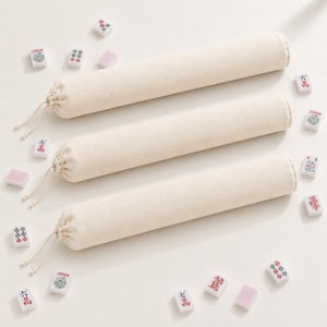 May include: Three long, cylindrical, cream-coloured fabric storage bags with drawstring closures at one end, arranged horizontally. Scattered around the bags are various rectangular mahjong tiles featuring colourful symbols, Chinese characters, and the word Joker.