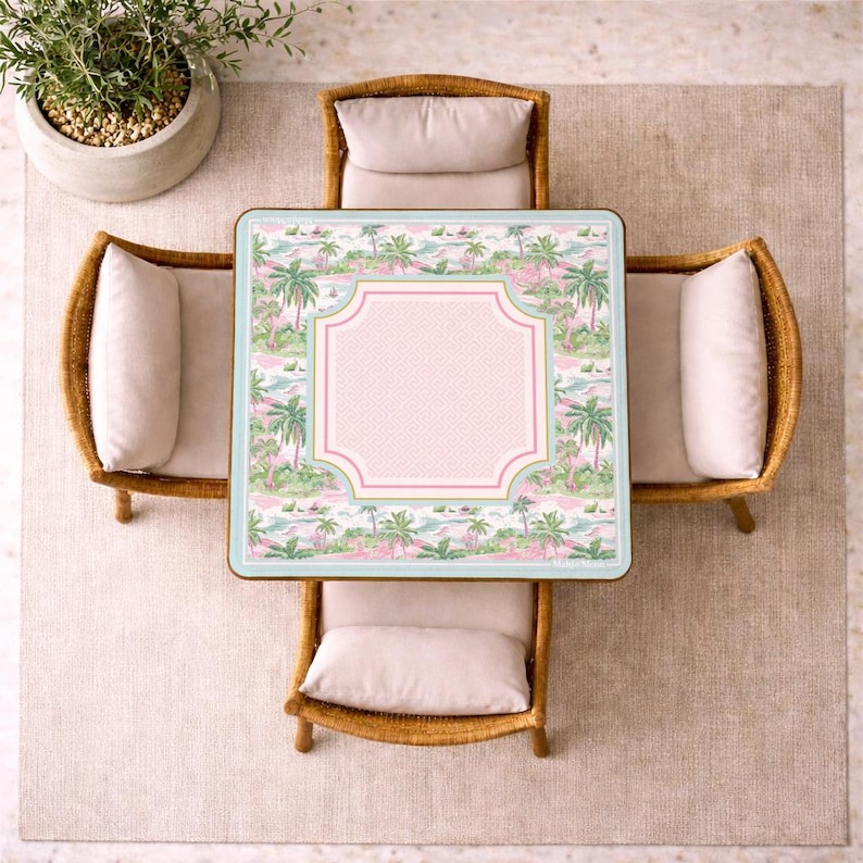 Op de afbeelding: Bovenaanzicht van een vierkante buitentafel met een tropisch landschapsontwerp, omringd door vier rotan stoelen met cr&egrave;mekleurige kussens. De tafel heeft een roze en wit patroon in het midden.