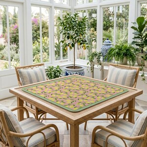 May include: A square table mat featuring a repeating pattern of yellow lemons and green leaves on a dusty pink background, bordered by a green and white checkered trim, centered on a light-colored wooden table with four matching chairs.