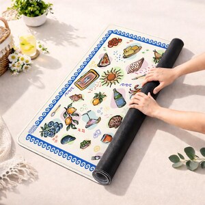 May include: A rectangular mat with a cream-colored surface featuring a blue wave-patterned border and various colorful illustrations including sardines, lemons, blueberries, a sun, and food items. The mat has a black underside and is being rolled up.