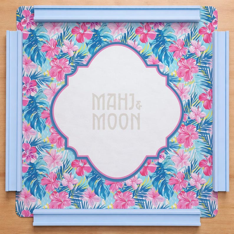 May include: A decorative tray with a floral pattern featuring pink hibiscus flowers and blue tropical leaves. The tray has a white centre with the text "MAHJ & MOON" and is framed by a light blue border.