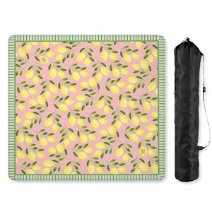 May include: A square picnic blanket featuring a repeating pattern of yellow lemons and green leaves on a light pink background, bordered by a green and white striped trim. A black cylindrical carrying bag with a shoulder strap is positioned nearby.