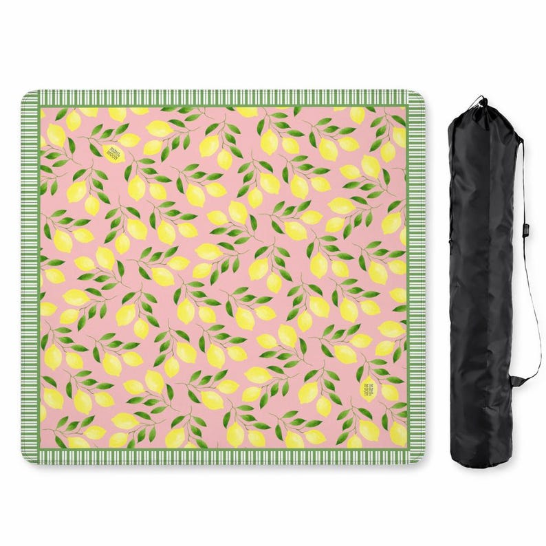May include: A square picnic blanket featuring a repeating pattern of yellow lemons and green leaves on a light pink background, bordered by a green and white striped trim. A black cylindrical carrying bag with a shoulder strap is positioned nearby.